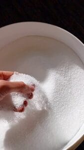 Hand schept witte kaarsenparels uit een kom – close-up van een hand die witte parelvormige kaarsenzand oppakt uit een ronde witte schaal. Het kaarsenzand heeft een fijne, korrelige structuur.