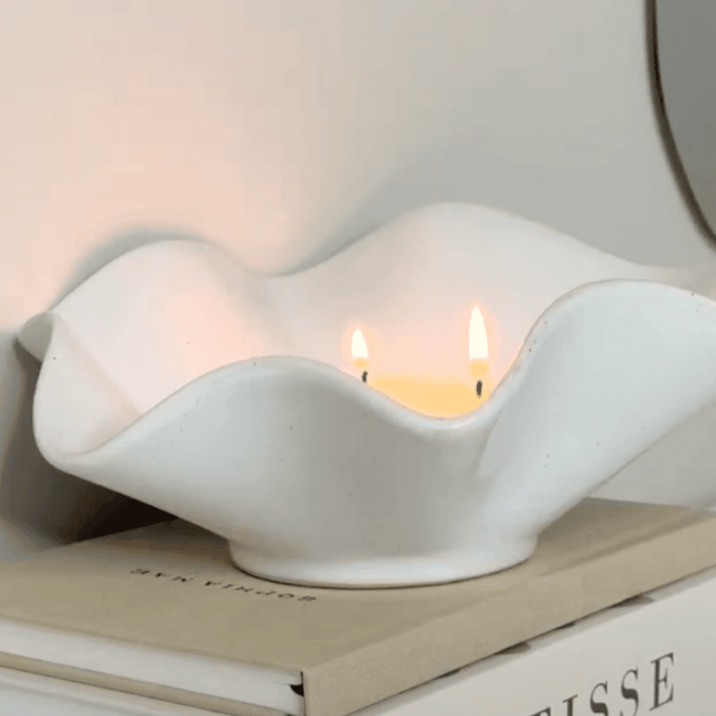 “Een witte keramische schaal met golvende randen, geplaatst op een stapel boeken. In de schaal brandt een kaars met twee vlammetjes, waardoor een zachte, warme gloed ontstaat. De vorm doet denken aan een open schelp, wat een elegante en rustgevende uitstraling geeft.”