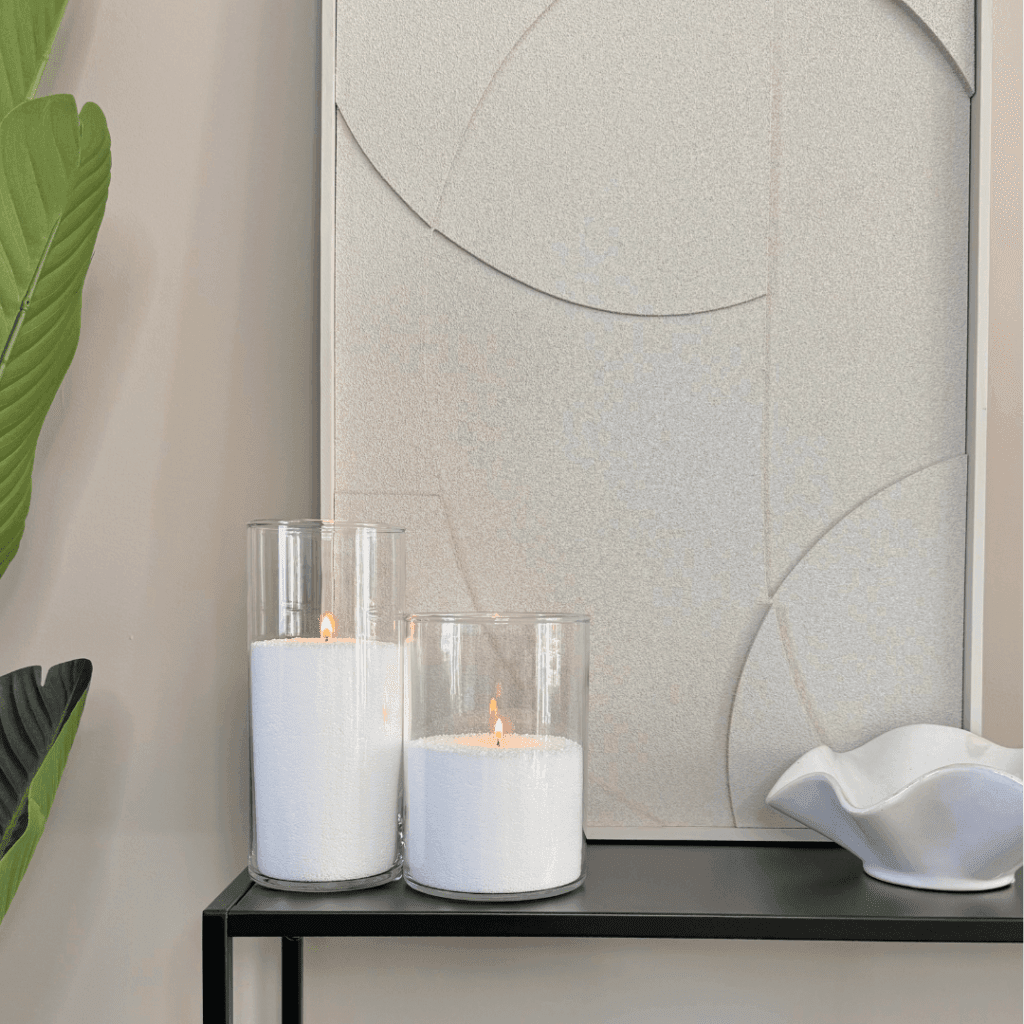 “Op een zwarte wandplank staan twee cilindervormige glazen vazen, gevuld met wit kaarsenzand. In elk zand is een kaarsje aangestoken, waardoor kleine vlammetjes branden. Rechts van de vazen staat een wit keramisch schaaltje met golvende randen, links is een groene plant zichtbaar. De achtergrond bestaat uit een minimalistisch reliefkunstwerk. Het geheel creëert een moderne, serene sfeer.”