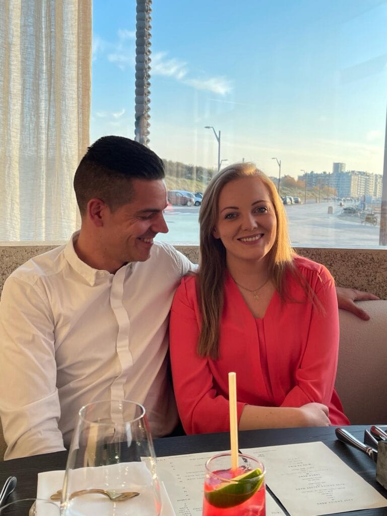 “Een man en een vrouw zitten samen in een zonnig restaurant. De man in een wit overhemd kijkt naar de vrouw, die een roze blouse draagt en glimlacht naar de camera. Op de tafel liggen menukaarten en staat een glas met een roze cocktail en een schijfje limoen. Door het grote raam zie je een heldere lucht, een boulevard en gebouwen in de verte. Het beeld straalt een warme, persoonlijke sfeer uit, passend bij de uitstraling van Pearl Candles.”