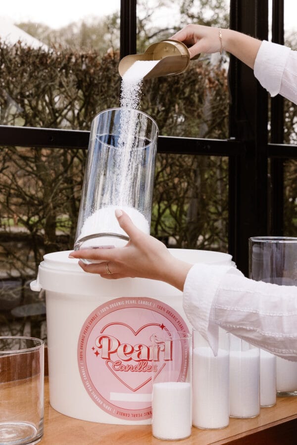 Kaarsenzand wordt zorgvuldig in een glazen houder gegoten vanuit een stijlvolle scoop, met meerdere gevulde glazen en een emmer op de achtergrond voor een elegante en praktische setting